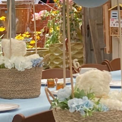 A decorated event table with wicker baskets filled with fluffy white and blue floral arrangements, yellow flowers in the background, place settings, and small honey jars as favors