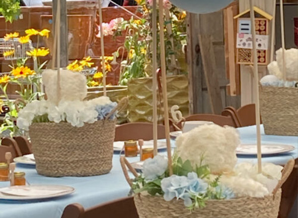 A decorated event table with wicker baskets filled with fluffy white and blue floral arrangements, yellow flowers in the background, place settings, and small honey jars as favors