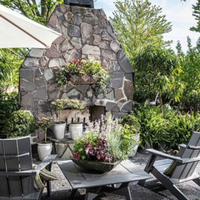 An outdoor garden patio with Adirondack chairs and a small table surrounded by lush potted plants and flowers, with a large stone fireplace feature in the background and a white patio umbrella