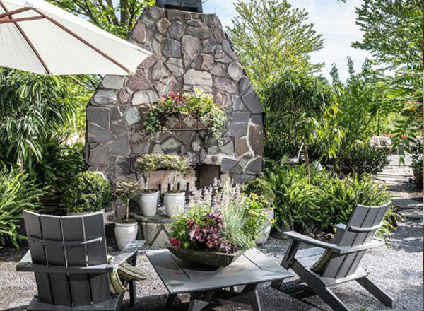 An outdoor garden patio with Adirondack chairs and a small table surrounded by lush potted plants and flowers, with a large stone fireplace feature in the background and a white patio umbrella
