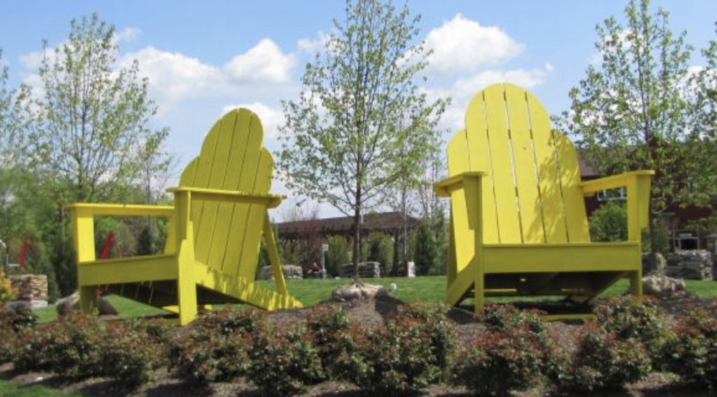 Two oversized yellow Adirondack chairs placed in a landscaped garden with shrubs and a green lawn under a blue sky with scattered clouds