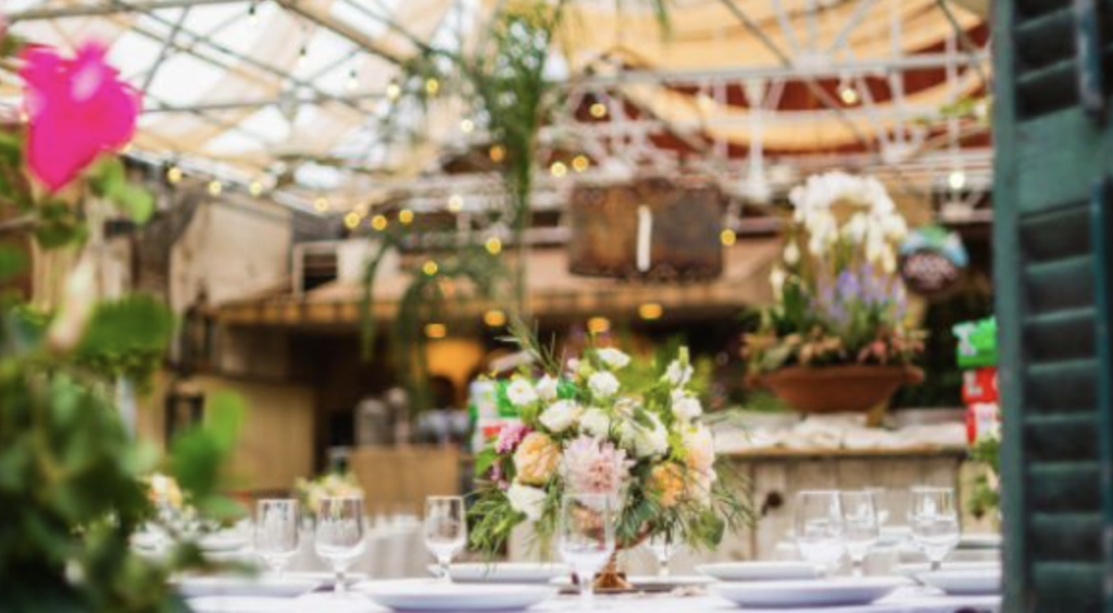 Elegant table setting with floral centerpiece in a greenhouse or garden venue, with glassware and plates arranged, blurred plants around