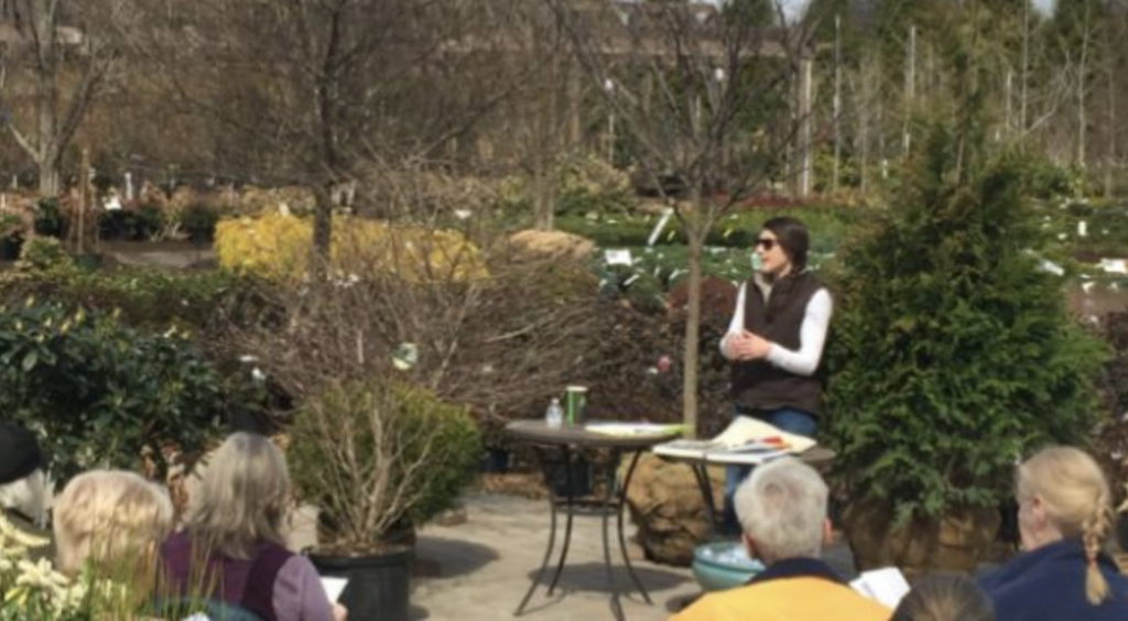 Outdoor garden presentation with a speaker standing beside a table addressing a seated audience, with shrubs and trees in the background