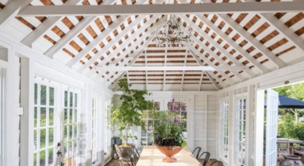Bright sunroom interior with white beams, glass windows, and potted plants on tables under a high ceiling with exposed rafters