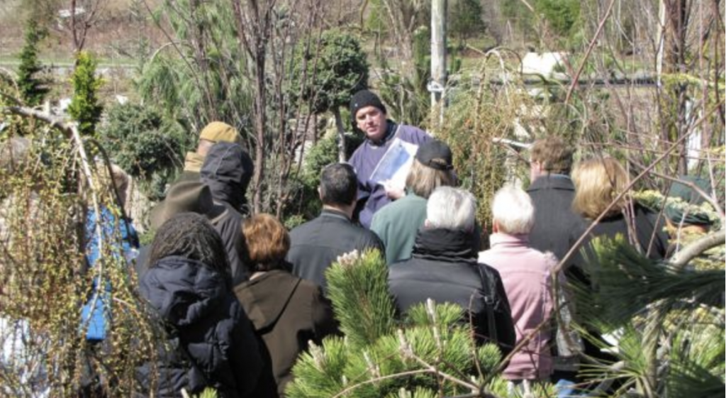 Group of people gathered outdoors around a presenter holding a binder during a garden demonstration among evergreen shrubs