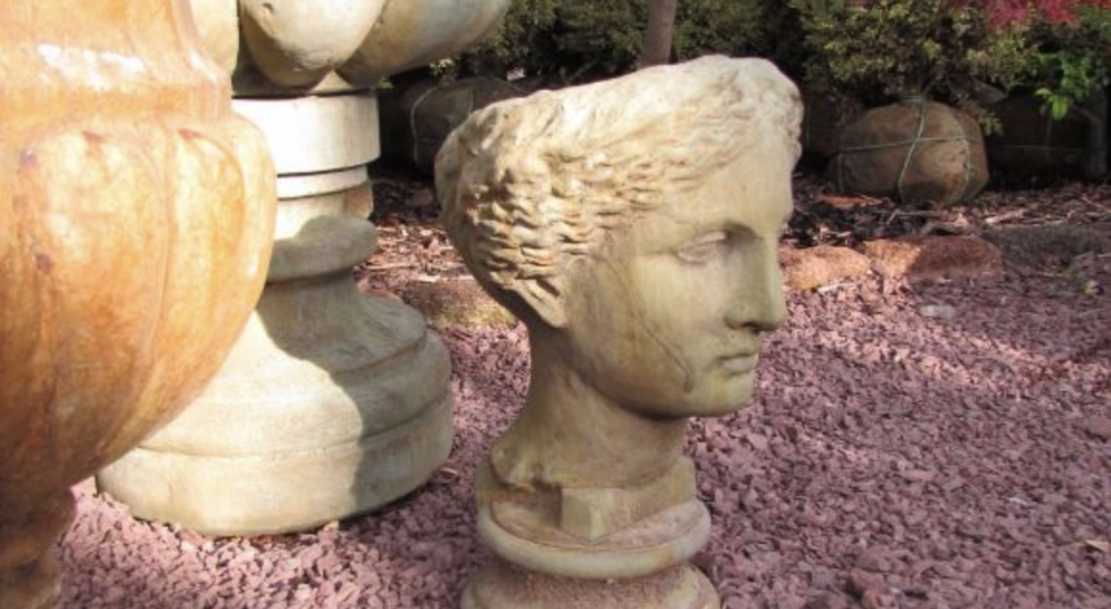 Stone bust planter featuring a classical face displayed among large round stone planters on a bed of red mulch.