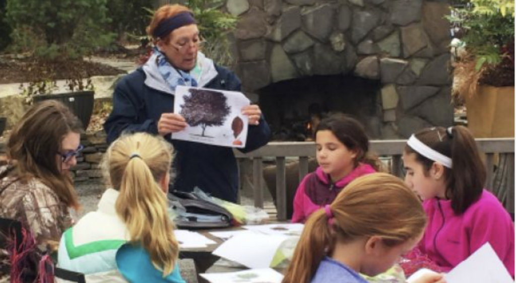 Adult instructor showing a picture of a tree to a group of young girls seated around a table outdoors during a plant education session.